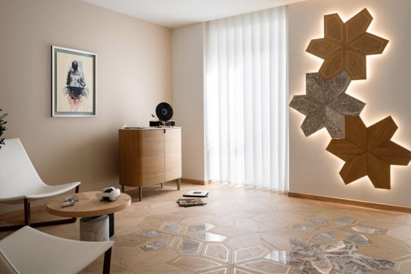 In this photo, you can see the lobby of a beauty and wellness center. The floor features the Azalea Module in Ca' Bassano light oak with a marble inlay. On the left, there are waiting chairs, while a painting hangs on the wall and a sideboard is positioned in the corner. In the background, a large window allows natural light into the space, and the Azalea 3D Art design with marble and brass inserts creates a striking decorative feature.