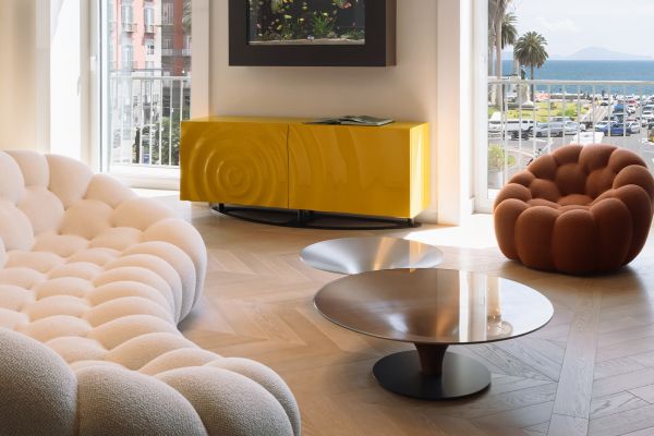 In this photo, you can see the living room of the Chevron 45° – Ca' Morosini – Napoli Apartment project. On the left, there is a sofa upholstered in white fabric that resembles a soft cloud. At the center of the room stands a glass coffee table, while on the right there is a small brick-colored armchair. In the background, beyond the large windows, a yellow sideboard supports a painting or television.