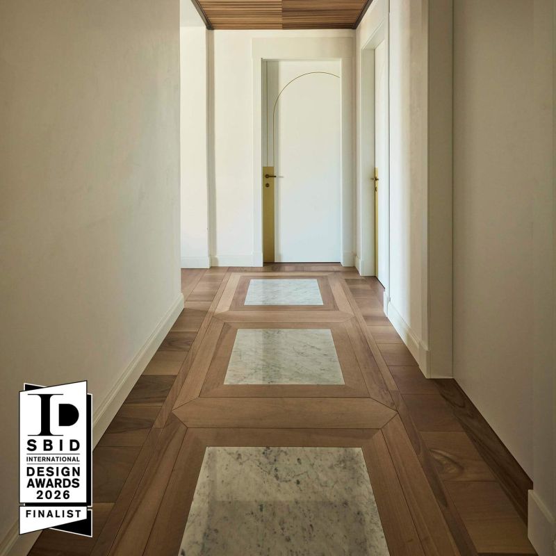 In this photo, you can see a corridor with white walls, a ceiling featuring Foglie d'Oro boiserie, and a floor that was selected as a finalist for the SBID 2026 Awards. The flooring is Matita by Fabio Rotella, installed in pattern 133, with an insect motif crafted in marble.
